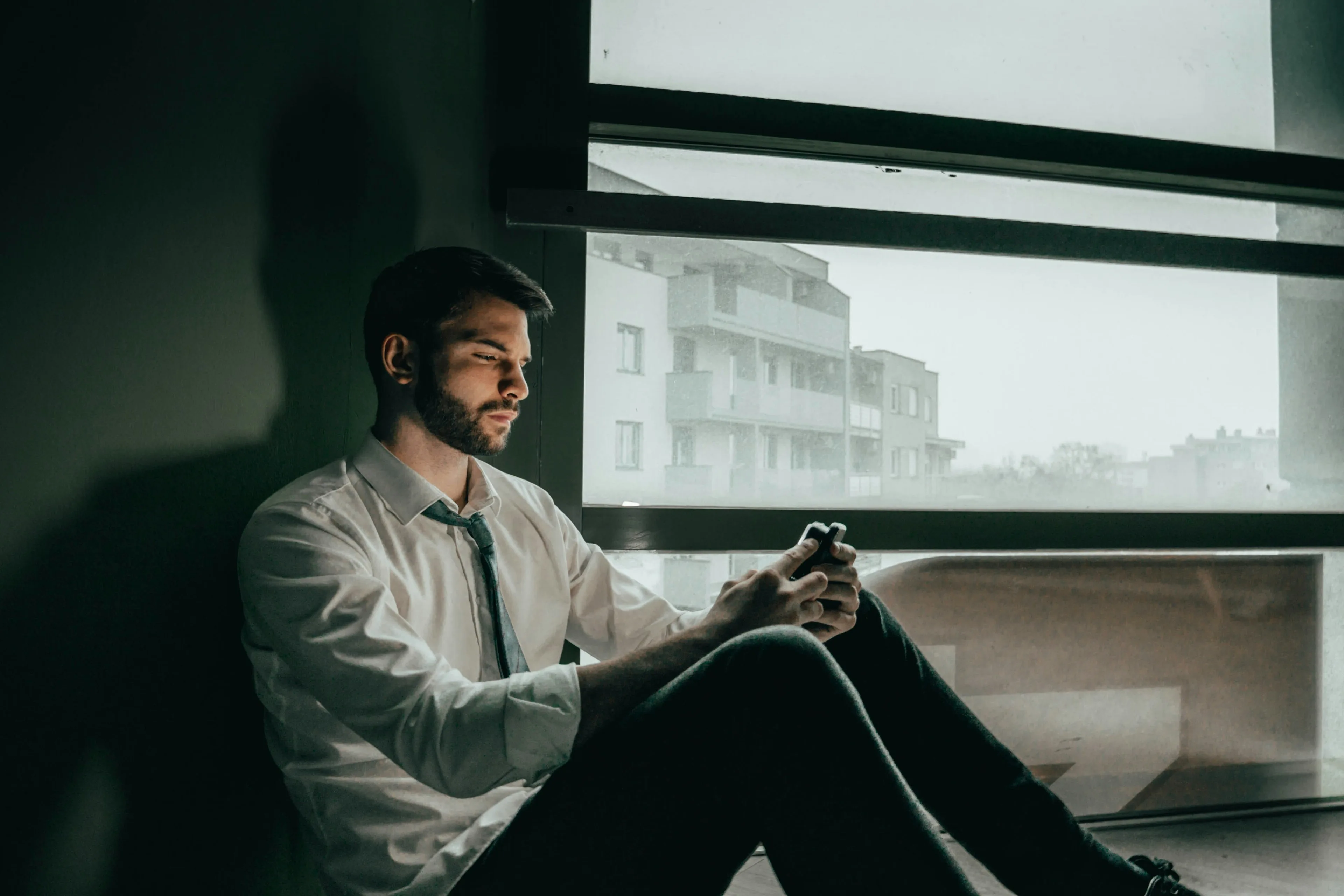A sad-looking man sitting on the floor and staring at his phone in an office