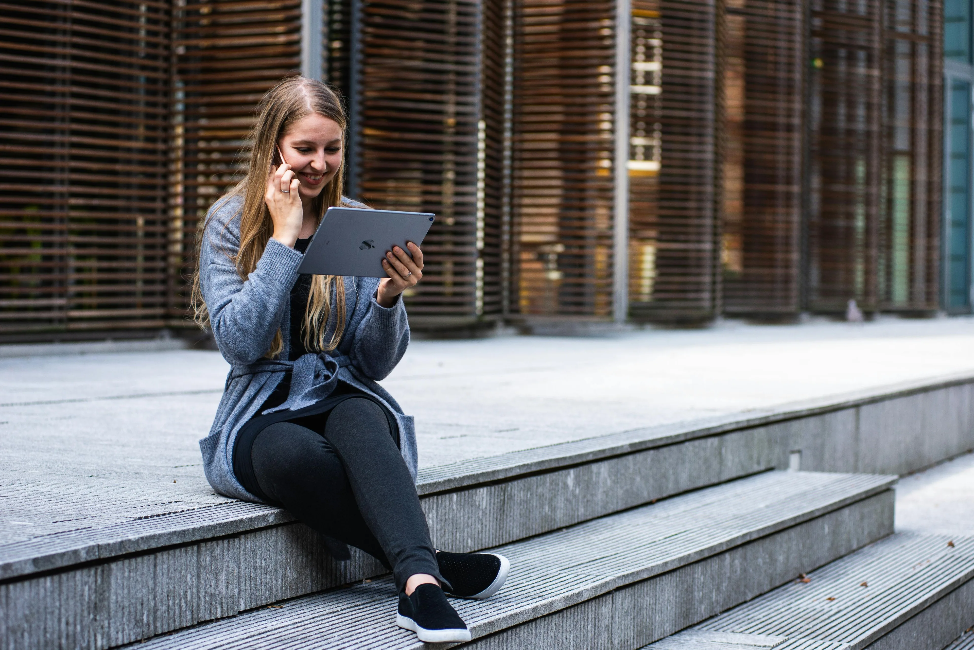 A happy-looking woman sitting on stairs outdoors, having a call while looking at a tablet.
